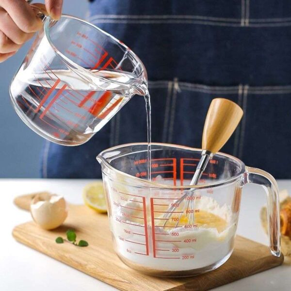 Female hand pouring water from a glass measuring cup into a mixture of flour and egg in another glass measuring cup.