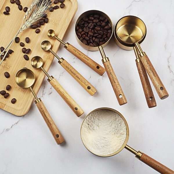 A picture showcasing four big golden measuring cups with wooden handles placed on a white marble table. One of the measuring cups contains coffee beans, while the second one holds flour. The remaining two cups are empty and stacked on top of each other. Additionally, there are four small measuring spoons with wooden handles arranged on a wooden board, which features scattered coffee beans and wheat.