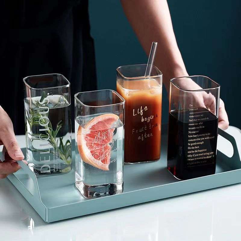 A thundercloud color serving tray on a table, showcasing four transparent drinking glasses. The first glass contains black coffee, the second glass holds a refreshing juice, the third glass features water with a slice of orange, and the fourth glass contains water infused with herbs.
