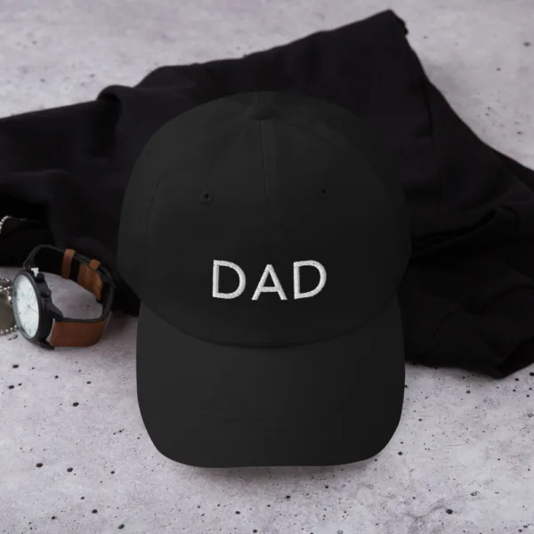 A black dad hat for Father's Day featuring white embroidered text 'DAD' on the front. The hat is placed on a black cloth with a brown leather watch beside it.