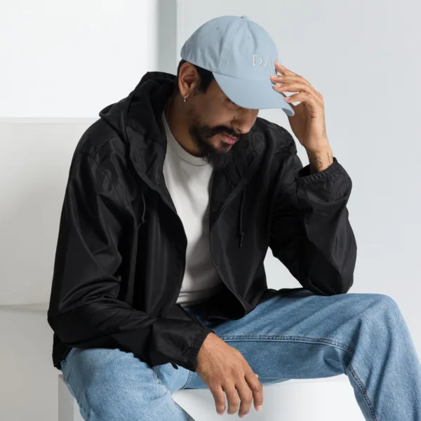 A man wearing a light blue dad hat for Father's Day with white embroidered text 'DAD' on the front. The man is sitting and looking down, gently touching the curved part of the dad hat.