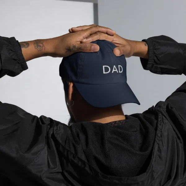 A man wearing a navy dad hat for Father's Day with white embroidered text 'DAD' on the front. The man is wearing the hat backwards and has his hands on top of his head, touching the hat.