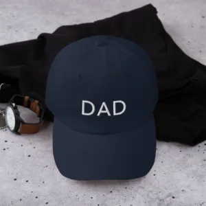 A navy dad hat for Father's Day featuring white embroidered text 'DAD' on the front. The hat is placed on a black cloth with a brown leather watch beside it.