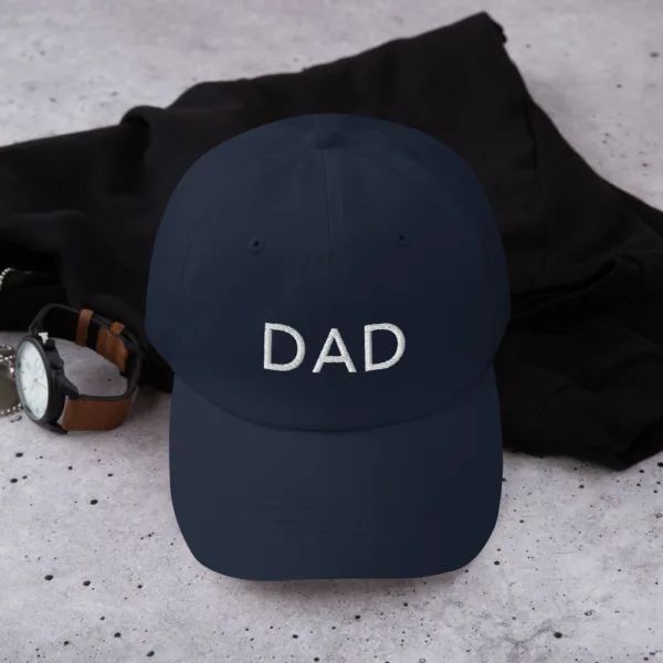 A navy dad hat for Father's Day featuring white embroidered text 'DAD' on the front. The hat is placed on a black cloth with a brown leather watch beside it.